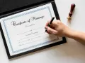 Dłoń pisze piórem na dokumencie "Certificate of Marriage", który poświadcza zawarcie związku małżeńskiego.