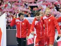 Trzech najlepszych piłkarzy ręcznych Polski w czerwonych strojach świętuje zwycięstwo. W tle kibice z biało-czerwonymi flagami.