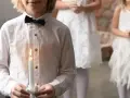 Dziecko w białej koszuli z czarną muszką trzyma zapaloną świecę. W tle inne dzieci w białych strojach.