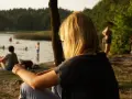 Dziewczyna z blond włosami siedzi nad jeziorem Trzechowskim, obserwując ludzi kąpiących się w wodzie.