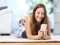 Uśmiechnięta dziewczyna w piżamie leży na dywanie, przeglądając telefon. Może szuka nowych znajomości na Tinder randki.