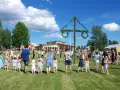Många människor, både vuxna och barn, håller varandra i händerna och dansar runt en midsommarstång. De firar midsommar i en grönskande park med gula byggnader i bakgrunden.