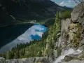 Widok z góry na Morskie Oko, odbijające chmury. Trasa turystyczna prowadzi przez skaliste zbocza z wodospadem.