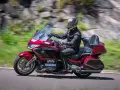 Mężczyzna w kasku jedzie lśniącym, czerwonym motocyklem uniwersalnym. W tle rozmyte zielone wzgórza i skały.