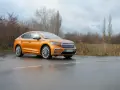 Pomarańczowa Skoda Enyaq Coupe na mokrej drodze, zarośla w tle.