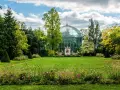Un jardín exuberante en el Bois de Boulogne, con una gran estructura de cristal verde y un cielo azul con nubes.