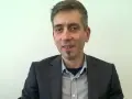 Maciej Kasprzak w czarnym garniturze i koszuli w kratę, z charakterystyczną bródką.