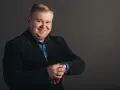 Uśmiechnięty mężczyzna w czarnym garniturze i niebieskiej koszuli, sprawdzający zegarek. Podpowiadamy, jak dobrać garnitur dla pana z brzuszkiem.