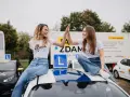 Dwie uśmiechnięte dziewczyny świętują sukces na dachu samochodu szkoleniowego. W tle ciężarówka z informacją o kursach na różne kategorie prawa jazdy.