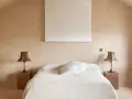 Camera da letto aesthetic con pareti color sabbia, letto bianco e due lampade in vimini. Un quadro astratto completa l'arredo.