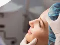 Kobieta w czepku medycznym, z rękami w rękawiczkach dotykającymi jej twarzy. Wygląda na to, że przeszła operację nosa, która nieudana.