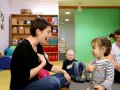 Eine Frau lacht und gestikuliert, während sie mit einem Kleinkind sprechen übt. Ein Junge und ein Mann sitzen im Hintergrund.