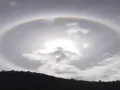 Un alone arcobaleno circonda il sole tra le nuvole, creando un effetto magico sopra una collina scura.