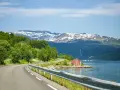 En slingrande väg längs en fjord i Norge, med snöklädda berg i bakgrunden. En röd stuga vid vattnet. Perfekt för en roadtrip norge rutt.