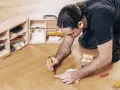 Mann repariert Parkett mit einem Reparaturstift und einer Farbauswahl. Werkzeugkasten mit Zubehör daneben.