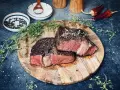 Zwei saftige Rib-Eye-Steaks, perfekt gegrillt, liegen auf einem Holzbrett. Mit Meersalz und Pfeffer bestreut, garniert mit frischem Thymian.