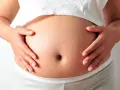 Brzuch ciężarnej kobiety, dłonie delikatnie go obejmują. Warto wiedzieć, jak rozpoznać sączące się wody płodowe.