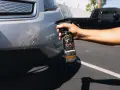 A hand sprays bug remover onto a car bumper, tackling stubborn bugs from a road trip.