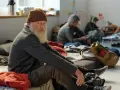 Älterer Mann mit Bart und Mütze sitzt auf dem Boden, umgeben von anderen Menschen. Eine Szene, die die Frage aufwirft: Wohin ohne Geld und Wohnung?