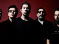 Pięciu mężczyzn, zespół Deftones Polska, pozuje do zdjęcia na ciemnoczerwonym tle.