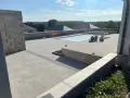 Vaste terrasse en béton gris avec piscine couverte, entourée de végétation et de maisons. Un chantier en cours.