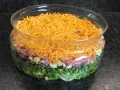 Une salade 7 étages colorée dans un grand bol en verre, avec des couches de laitue, petits pois, oignons rouges, jambon, œufs durs et fromage râpé.