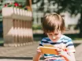 Chłopiec w kolorowym T-shircie ogląda film dla małych dzieci na żółtym telefonie, siedząc na zewnątrz.