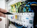 Ręka majstruje przy bezpiecznikach. Kolorowe przewody tworzą skomplikowaną instalację elektryczną.