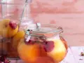 Zwei Gläser mit eingelegten Pfirsichen, verziert mit Rosenblättern und Gewürzen, auf einem rosa Holztisch.