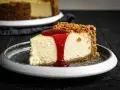 Ein Stück cremiger american cheesecake mit Keksboden, übergossen mit roter Fruchtsauce, auf einem grauen Teller.