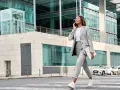 Elegancki strój casual damski: kobieta w szarym garniturze i białej koszulce rozmawia przez telefon, niosąc laptopa.