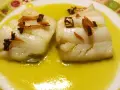 Dos trozos de bacalao al pil pil, cubiertos con ajo frito y guindilla, nadan en una salsa dorada sobre un plato decorado.