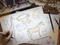 Un dessin d'animaux réaliste facile : une main esquisse une girafe, entourée de croquis de corgis, d'un cheval, d'un iguane, d'un requin et d'un oiseau.