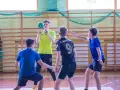 Cezary Surgiel w żółtej koszulce rzuca piłkę do ręcznej, otoczony przez kolegów z drużyny na sali gimnastycznej.