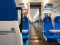 Wnętrze pociągu TLK: rzędy niebieskich siedzeń z białymi zagłówkami i stolikami. Widać przejście i ekrany informacyjne.