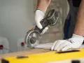 Ręce w białych rękawiczkach trzymają szlifierkę kątową, precyzyjnie tnąc płytkę. To pokazuje, jak ciąć płytki szlifierką kątową.