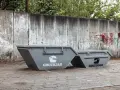 Dwa szare kontenery na śmieci zmieszane, z logo GRUZOJAD, stoją przed zniszczonym betonowym murem pokrytym graffiti.