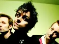 Trzej członkowie Green Day: Billie Joe Armstrong, Mike Dirnt i Tré Cool, pozują do zdjęcia na zielonym tle.