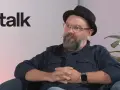 Mężczyzna w okularach i kapeluszu siedzi, rozmawiając. Obok logo DGP talk.