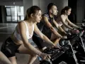 Trzy osoby ćwiczą na rowerach stacjonarnych. Kobieta z przodu w białym topie i czarnych spodenkach, obok mężczyzna w słuchawkach i kobieta w różowym topie. To świetny sposób na trening FBW.