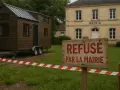 Une tiny house sur roues est devant la mairie. Un panneau indique "Refusé par la mairie", barré par un ruban de sécurité.