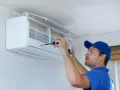Un technicien en bleu utilise un tournevis pour ajuster un climatiseur mural, assurant le bon fonctionnement pour recharger clim maison.