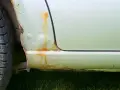 Rdza i pęcherze na nadkolu samochodu, obok opony. Widoczne zacieki rdzy na jasnej karoserii.