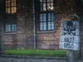 Tablica z symbolem czaszki i piszczeli oraz napisem "HALT! STÓJ!" na tle ceglanego budynku i drutu kolczastego, przypominająca o grozie obozów koncentracyjnych.