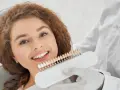 Uśmiechnięta kobieta z rudymi włosami porównuje odcień swoich zębów z paletą kolorów. Dowiedz się, jak mieć białe zęby bez wybielania.