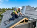 Fachowiec w kasku i kamizelce wykonuje obróbkę blacharską dachu, przykręcając metalowe elementy.
