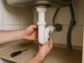 Mains de personne réparant un siphon bouché sous un évier. Un tournevis est posé sur une étagère.