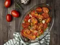Pomodori gratinati al forno, dorati e succosi, con cipolle e rosmarino in una teglia di vetro su un tavolo di legno.