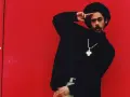Mann mit Afro und Schnurrbart, schwarzer Rollkragenpullover, goldene Kette mit Kreuzanhänger, vor rotem Hintergrund. Er salutiert. "I shot the sheriff" könnte man sagen.