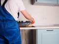 Instalator w niebieskim kombinezonie podłącza kuchenkę gazową w kuchni.